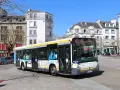 Un bus Heuliez GX 327 blanc et jaune circule dans une rue pavée, devant des immeubles anciens sous un ciel bleu.
