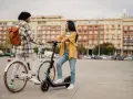 Deux jeunes femmes discutent, l'une sur un vélo blanc, l'autre sur une trottinette électrique. Elles profitent d'un moyen de transport doux en ville.