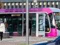 Un tramway rose et gris, un des transports en commun de Metz, s'arrête à un arrêt. Des passagers montent et descendent.