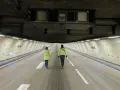 Deux personnes en gilets jaunes traversent un tunnel duplex de l'A86, la hauteur impressionnante du plafond est soulignée par les équipements techniques.