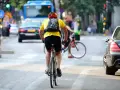 Un cycliste en tenue jaune et casque rouge profite de faire du vélo en ville, traversant une rue animée avec des voitures et un bus.