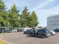 Un homme en costume monte dans un SUV gris dans un parking. Des bus électriques sont garés à proximité, près d'un bâtiment moderne. C'est une scène typique de naolib Nantes.
