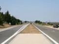 Une longue voie publique s'étend sous un ciel bleu clair. Des arbres bordent la route, et une voiture est garée sur le bas-côté.