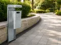 Poubelle métallique sur une voie urbaine bordée de verdure et d'un mur de pierre.