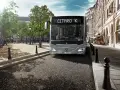 Un bus Mercedes-Benz Citaro K gris, dont le poids est impressionnant, roule dans une rue pavée bordée de bâtiments haussmanniens et d'arbres.