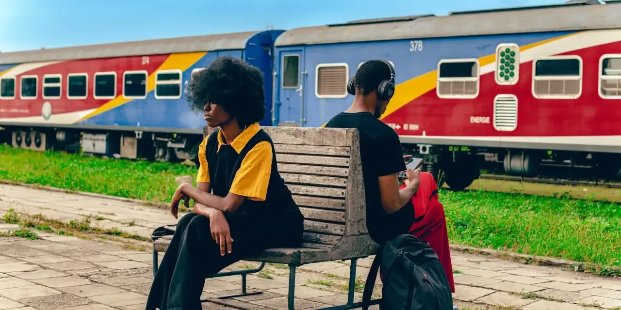 Deux personnes attendent sur un banc, un train coloré, un moyen de transport en Afrique, stationne derrière elles.