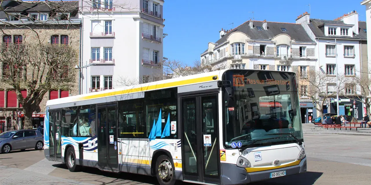 Un bus Heuliez GX 327 blanc et jaune circule dans une rue pavée, devant des immeubles anciens sous un ciel bleu.
