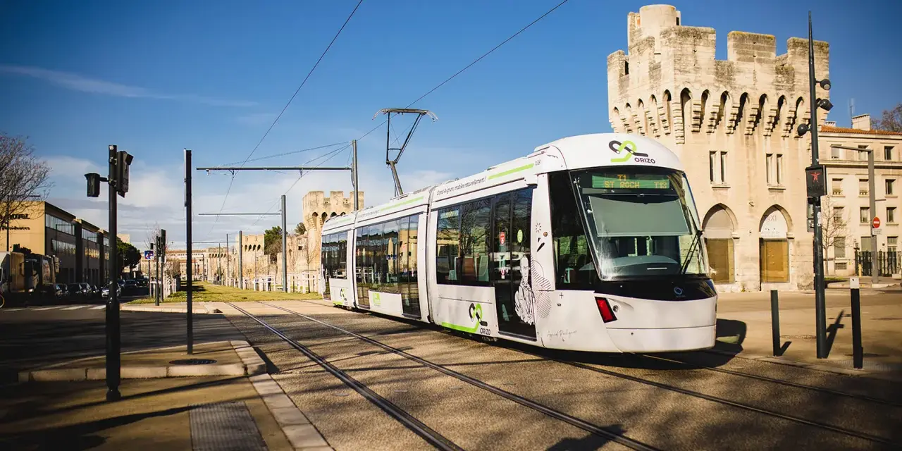 Le tramway moderne circule sur les rails à Avignon, passant devant des bâtiments historiques sous un ciel bleu éclatant.