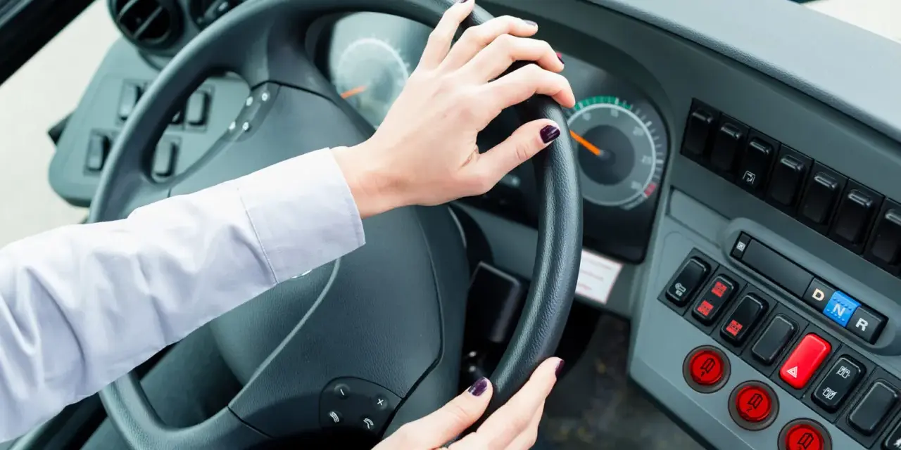 Vue de l'intérieur d'un bus, mains sur le volant. Le tableau de bord est visible, avec des boutons et des indicateurs. Le chauffeur est prêt à partir.