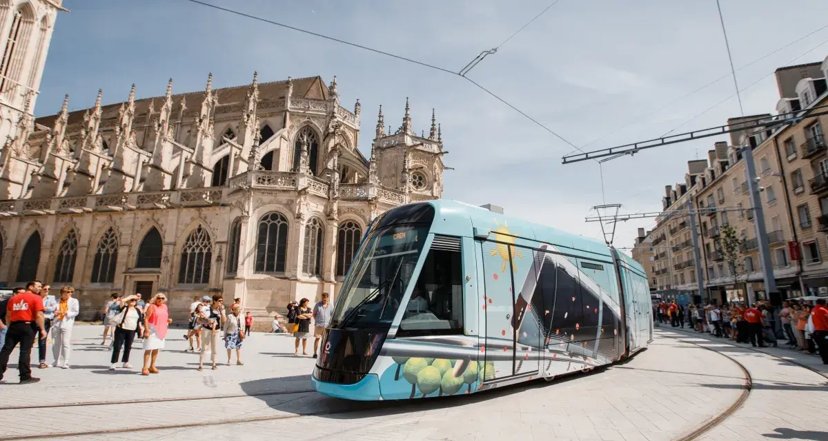 Un tram **Keolis Vélizy** décoré circule devant une cathédrale gothique. Des passants attendent sur le quai.