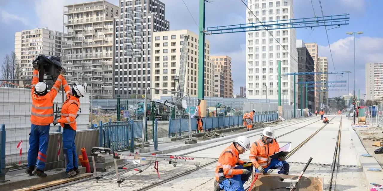 Des ouvriers travaillent sur de nouvelles infrastructures de tramway, avec des immeubles modernes en arrière-plan.