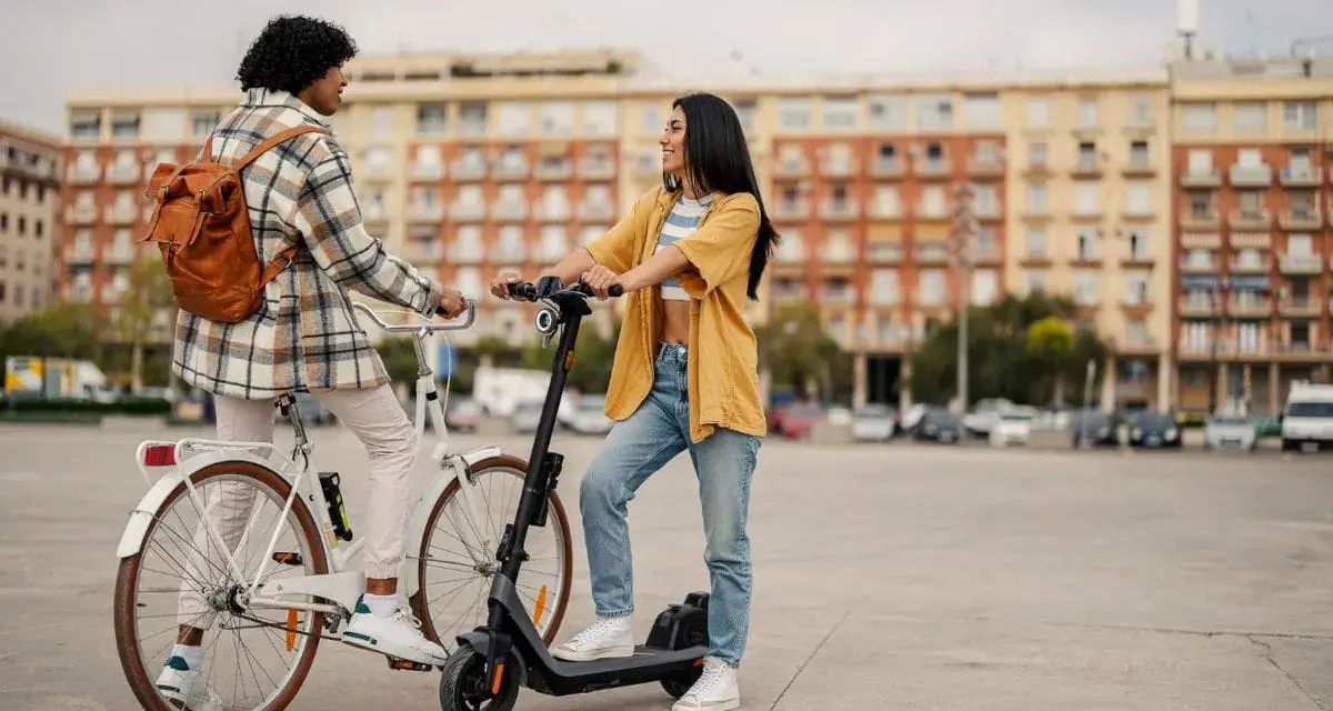 Deux jeunes femmes discutent, l'une sur un vélo blanc, l'autre sur une trottinette électrique. Elles profitent d'un moyen de transport doux en ville.