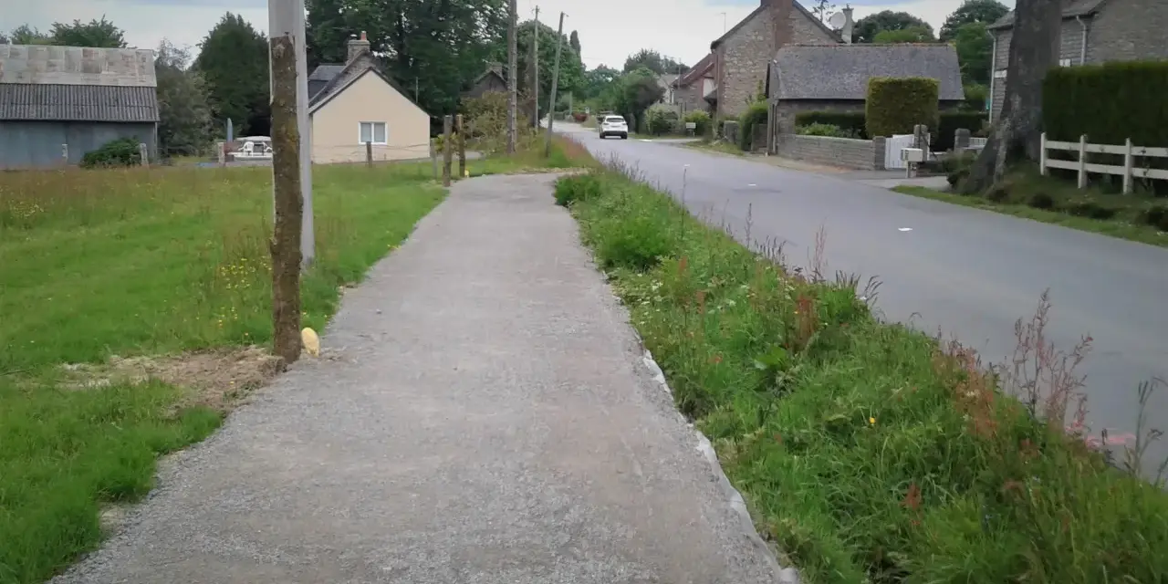 Un chemin de gravier longe une route de campagne bordée d'herbes folles. C'est un exemple de cheminement doux, invitant à la promenade paisible.
