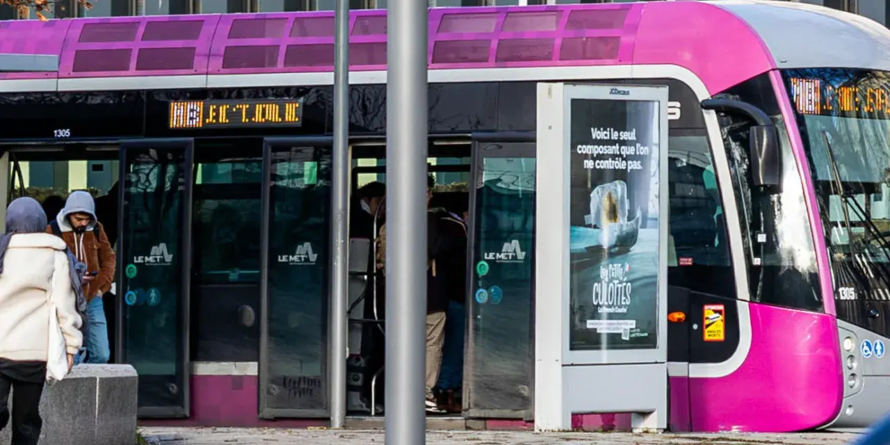 Un tramway rose et gris, un des transports en commun de Metz, s'arrête à un arrêt. Des passagers montent et descendent.