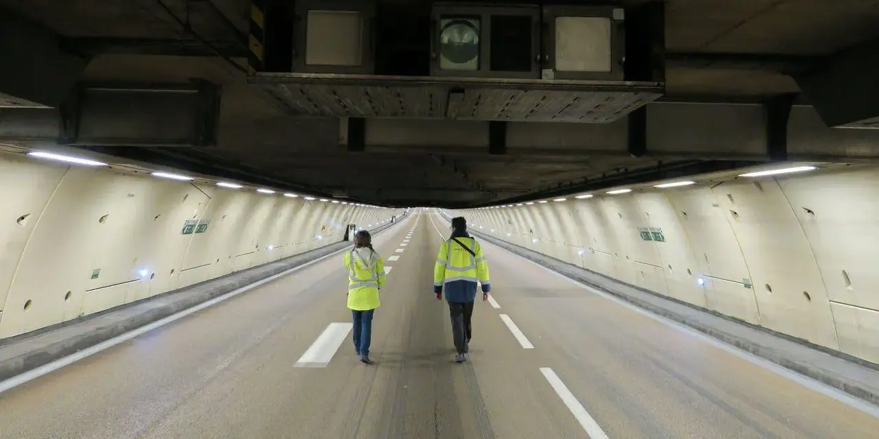 Deux personnes en gilets jaunes traversent un tunnel duplex de l'A86, la hauteur impressionnante du plafond est soulignée par les équipements techniques.