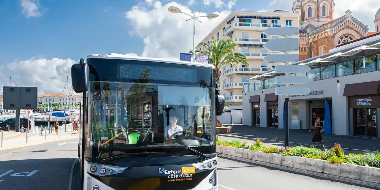 Un bus Esterel côte d'Azur circule dans une ville ensoleillée, près d'un port et d'une église.