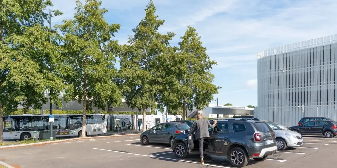 Un homme en costume monte dans un SUV gris dans un parking. Des bus électriques sont garés à proximité, près d'un bâtiment moderne. C'est une scène typique de naolib Nantes.
