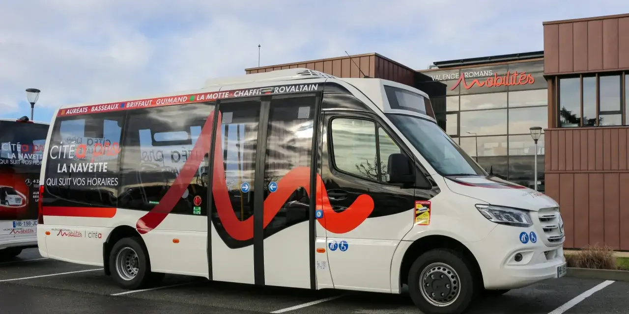 Un bus blanc avec des motifs rouges et noirs, portant l'inscription "Citéa La Navette" et des noms de localités, est garé devant un bâtiment "Valence Romans Mobilités".