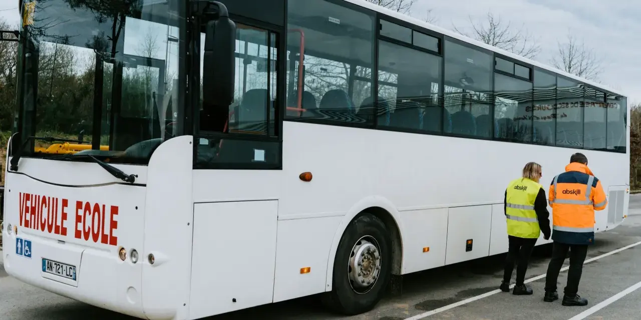 Un bus blanc "Véhicule École" est garé. Deux instructeurs, un homme et une femme, portent des gilets de haute visibilité.