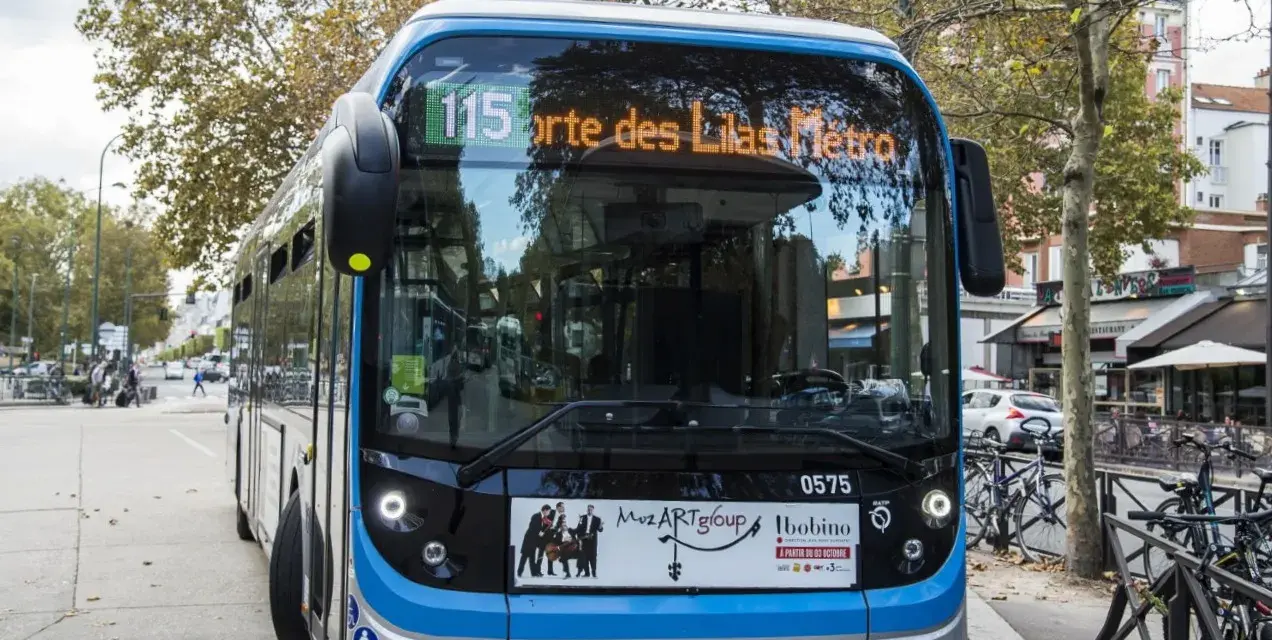 Bus Bolloré 115, direction Porte des Lilas Métro, stationné dans une rue bordée d'arbres et de commerces.