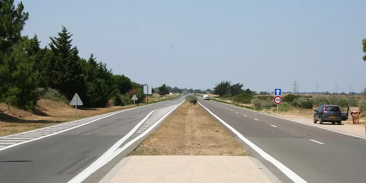 Une longue voie publique s'étend sous un ciel bleu clair. Des arbres bordent la route, et une voiture est garée sur le bas-côté.