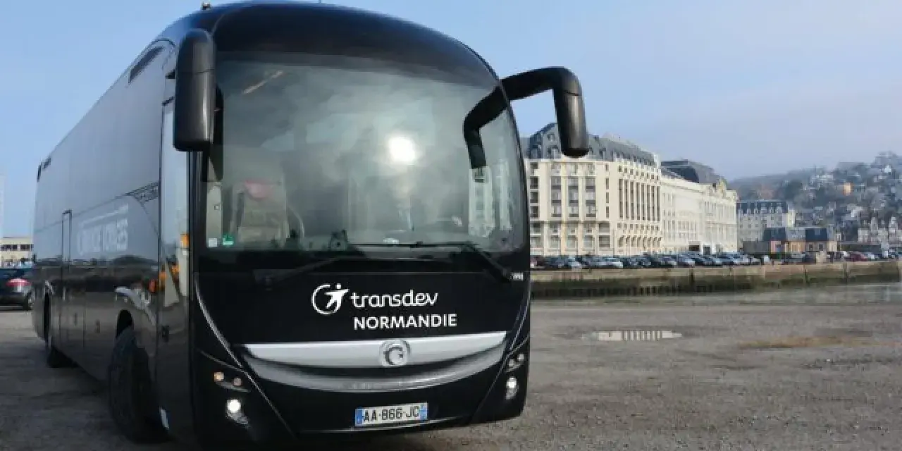 Autocar noir Transdev Normandie garé sur un quai, avec des bâtiments en arrière-plan.
