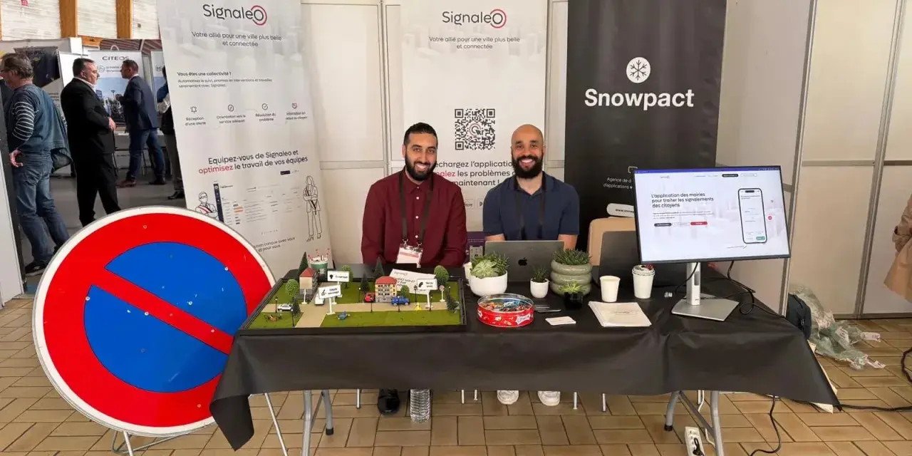 Deux hommes souriants présentent une maquette de ville et un panneau "Interdit de stationner" pour illustrer leur solution de **service voirie** connectée.