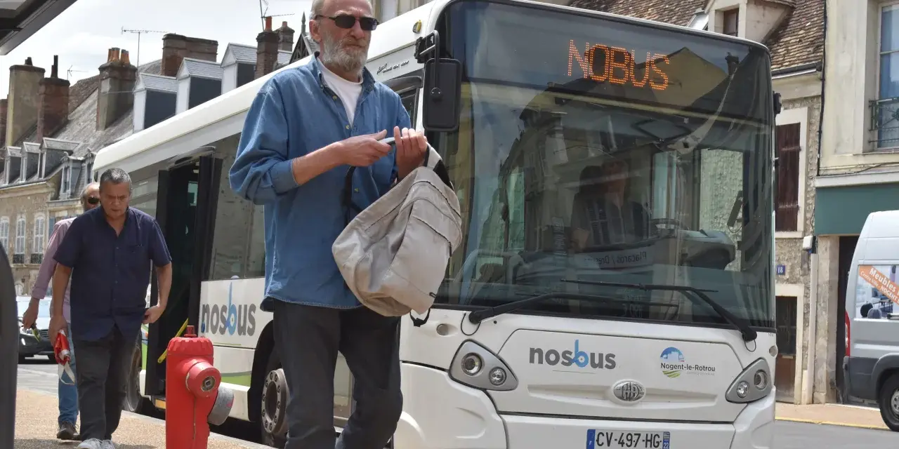 Un homme attend le bus à Nogent-le-Rotrou, sac à la main. Le panneau lumineux indique "NOBUS".