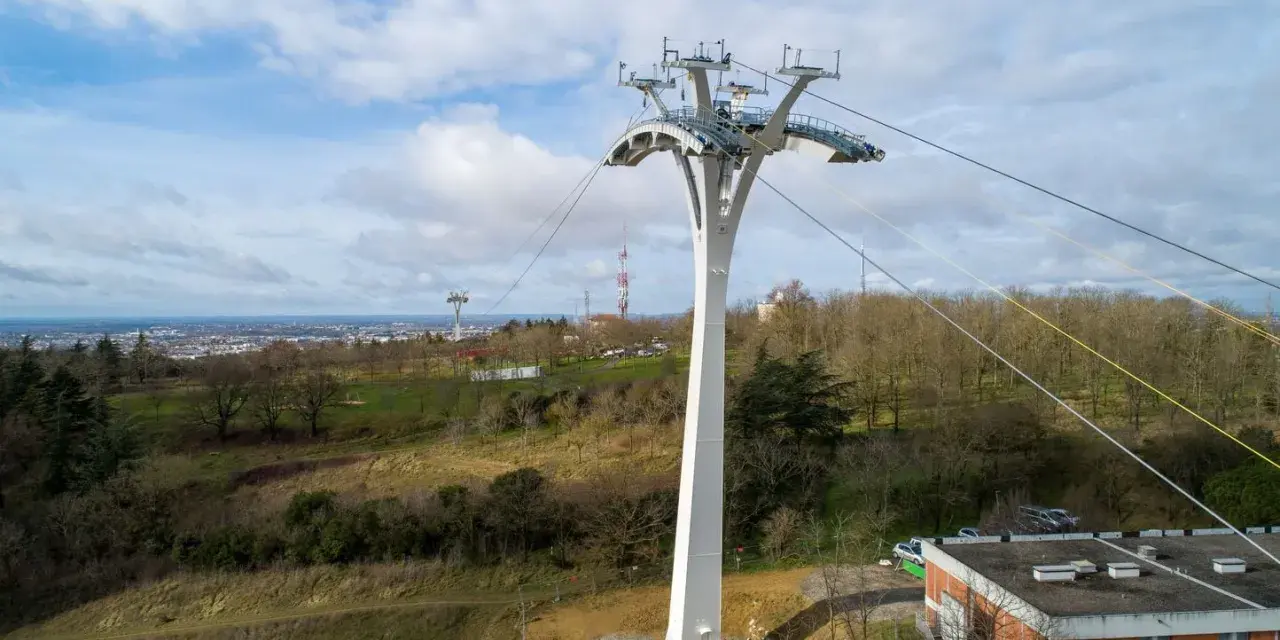 Un pylône blanc massif soutient un téléphérique urbain au-dessus d'une ville et d'une colline boisée.