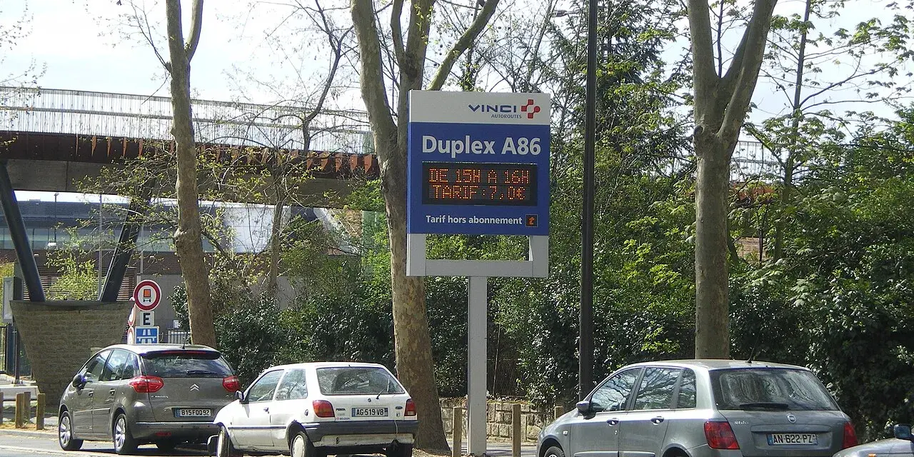 Panneau lumineux Vinci Autoroutes pour le Duplex A86, indiquant les tarifs. Des voitures sont garées sur le bord de la route.