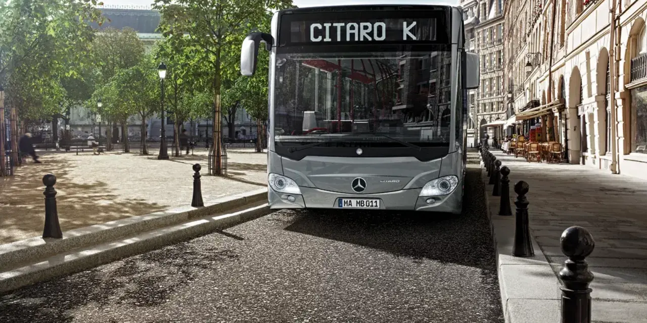 Un bus Mercedes-Benz Citaro K gris, dont le poids est impressionnant, roule dans une rue pavée bordée de bâtiments haussmanniens et d'arbres.