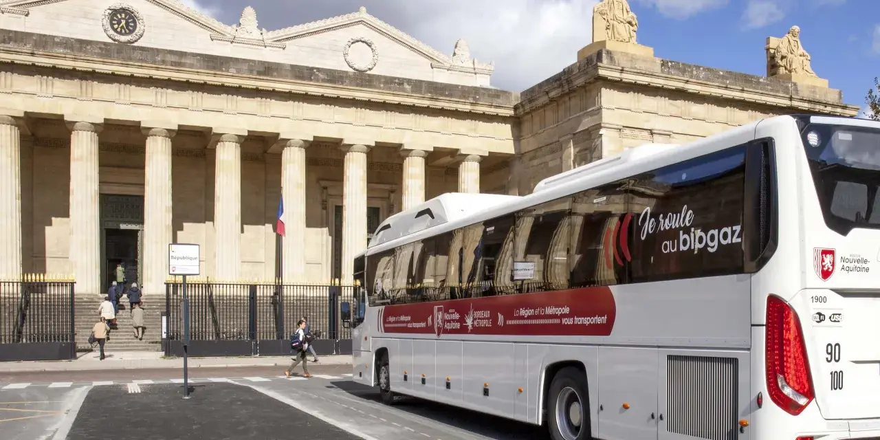 Un bus blanc "car régionaux Gironde" roule devant un bâtiment imposant avec des colonnes. Le bus arbore le slogan "Je roule au bipgaz".