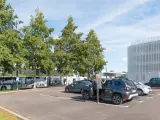 Un homme en costume monte dans un SUV gris dans un parking. Des bus électriques sont garés à proximité, près d'un bâtiment moderne. C'est une scène typique de naolib Nantes.