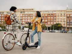 Deux jeunes femmes discutent, l'une sur un vélo blanc, l'autre sur une trottinette électrique. Elles profitent d'un moyen de transport doux en ville.