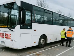 Un bus blanc "Véhicule École" est garé. Deux instructeurs, un homme et une femme, portent des gilets de haute visibilité.