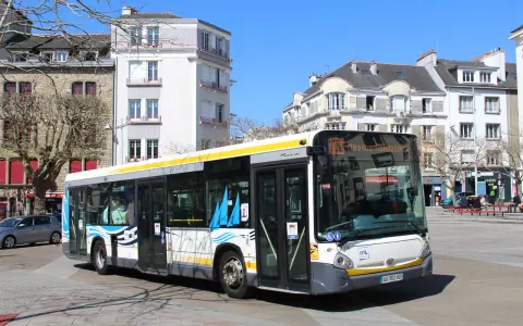 Un bus Heuliez GX 327 blanc et jaune circule dans une rue pavée, devant des immeubles anciens sous un ciel bleu.