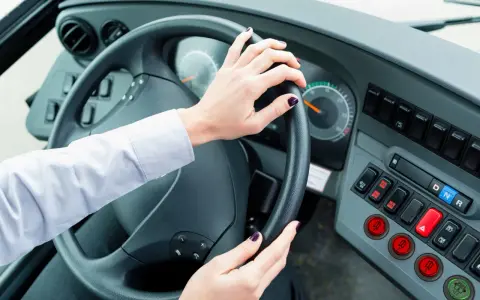 Vue de l'intérieur d'un bus, mains sur le volant. Le tableau de bord est visible, avec des boutons et des indicateurs. Le chauffeur est prêt à partir.