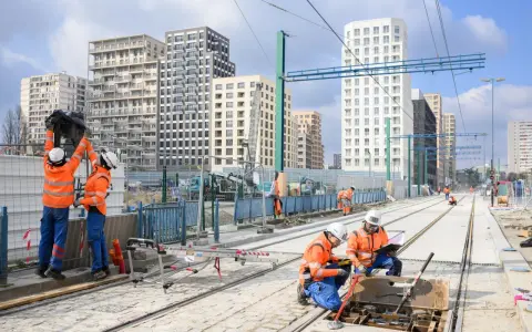 Des ouvriers travaillent sur de nouvelles infrastructures de tramway, avec des immeubles modernes en arrière-plan.