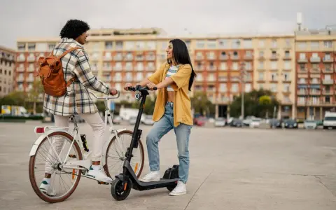 Deux jeunes femmes discutent, l'une sur un vélo blanc, l'autre sur une trottinette électrique. Elles profitent d'un moyen de transport doux en ville.