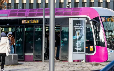 Un tramway rose et gris, un des transports en commun de Metz, s'arrête à un arrêt. Des passagers montent et descendent.