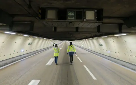 Deux personnes en gilets jaunes traversent un tunnel duplex de l'A86, la hauteur impressionnante du plafond est soulignée par les équipements techniques.