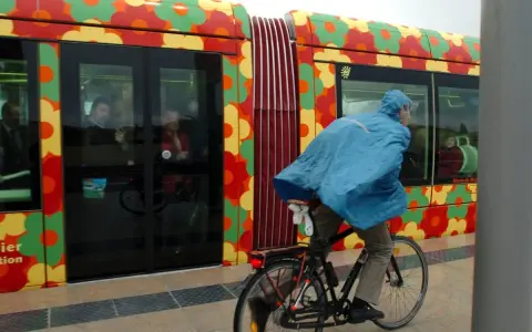 Cycliste sous la pluie, longeant un tramway décoré de fleurs colorées à Montpellier.