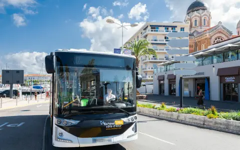 Un bus Esterel côte d'Azur circule dans une ville ensoleillée, près d'un port et d'une église.