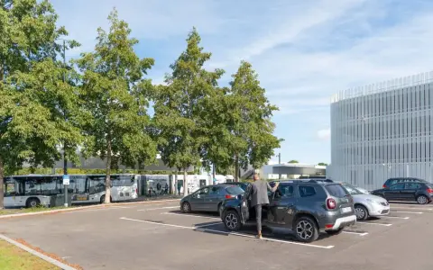 Un homme en costume monte dans un SUV gris dans un parking. Des bus électriques sont garés à proximité, près d'un bâtiment moderne. C'est une scène typique de naolib Nantes.