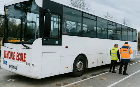 Un bus blanc "Véhicule École" est garé. Deux instructeurs, un homme et une femme, portent des gilets de haute visibilité.