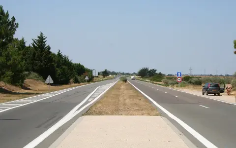 Une longue voie publique s'étend sous un ciel bleu clair. Des arbres bordent la route, et une voiture est garée sur le bas-côté.