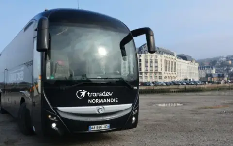 Autocar noir Transdev Normandie garé sur un quai, avec des bâtiments en arrière-plan.