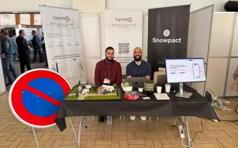 Deux hommes souriants présentent une maquette de ville et un panneau "Interdit de stationner" pour illustrer leur solution de **service voirie** connectée.