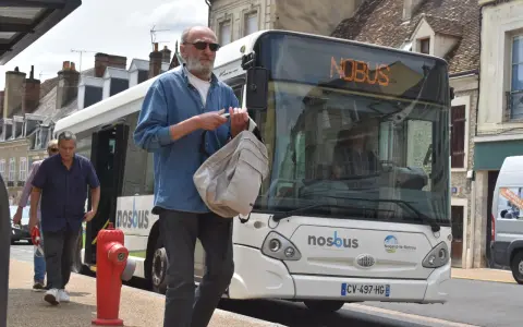 Un homme attend le bus à Nogent-le-Rotrou, sac à la main. Le panneau lumineux indique "NOBUS".