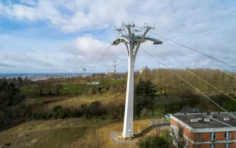 Un pylône blanc massif soutient un téléphérique urbain au-dessus d'une ville et d'une colline boisée.